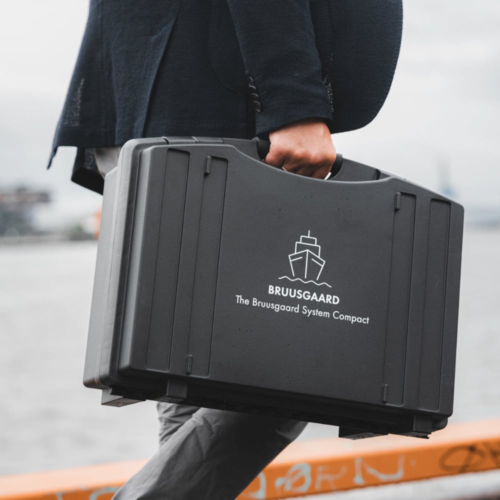 Person carrying a black Bruusgaard case labeled ‘The Bruusgaard System Compact’ by the water.